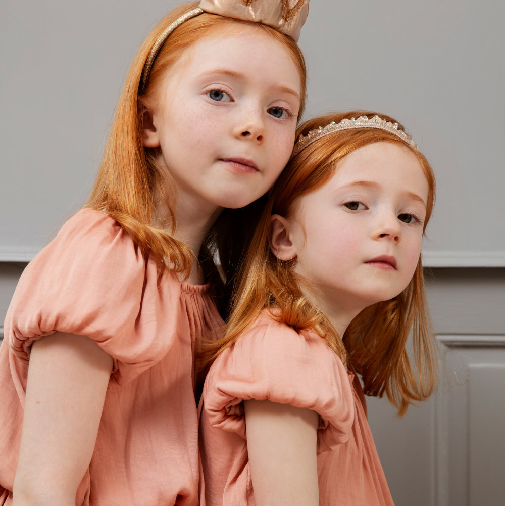 Two young girls in matching pink dresses with headbands, sitting close together.