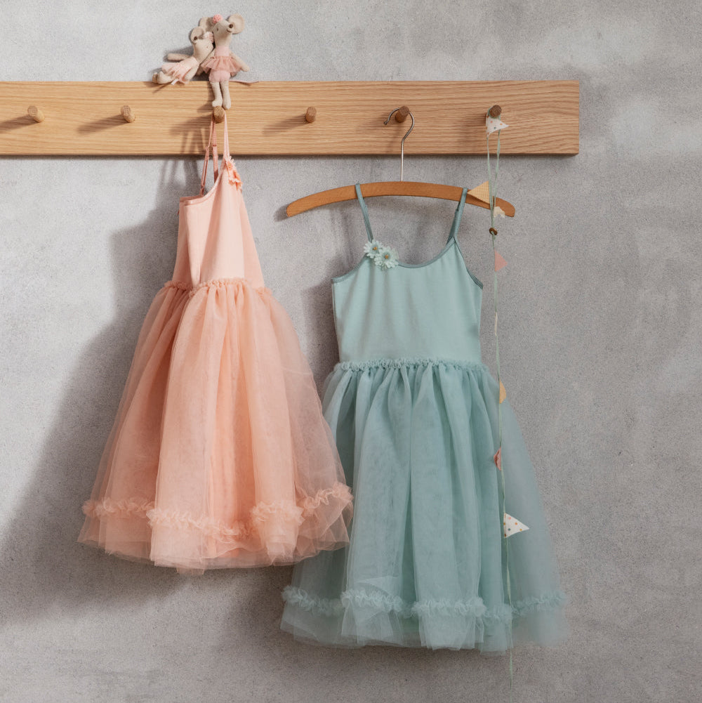 Two children's dresses, one pink and one green, hanging on a wooden rack against a gray wall.