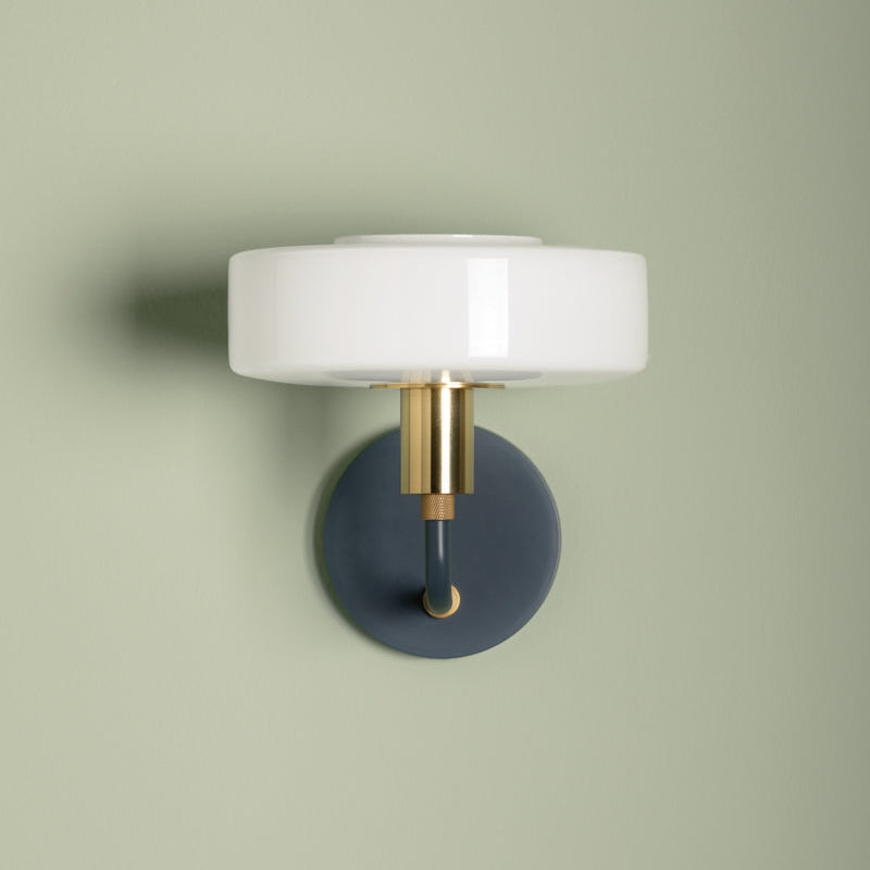 Wall-mounted light fixture with white shade and gold and blue base on a green wall