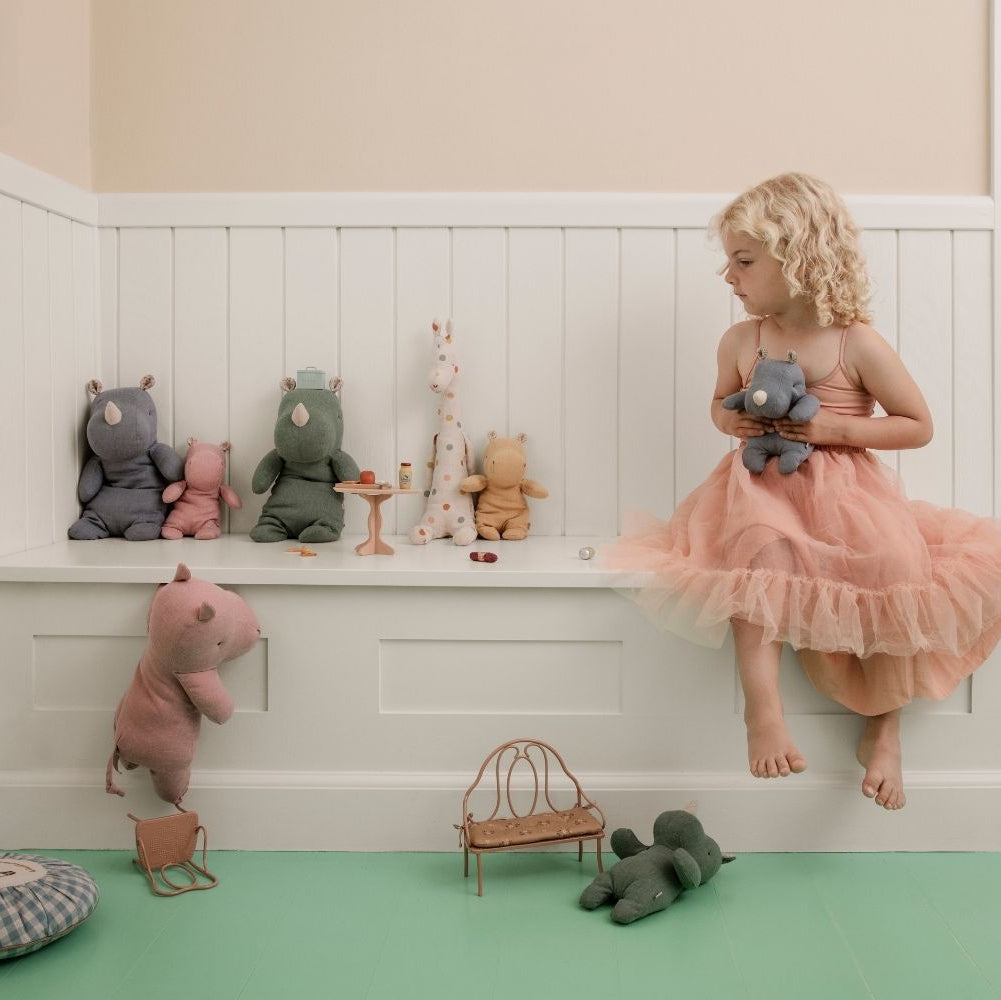 Child in a pink dress sitting on a white shelf with stuffed animals in a room with green floor and beige walls.