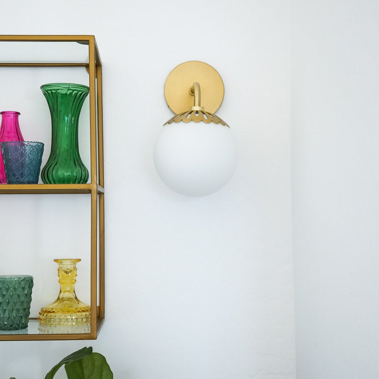 Wall sconce with a white globe shade next to a gold shelf with colorful vases on a white wall.