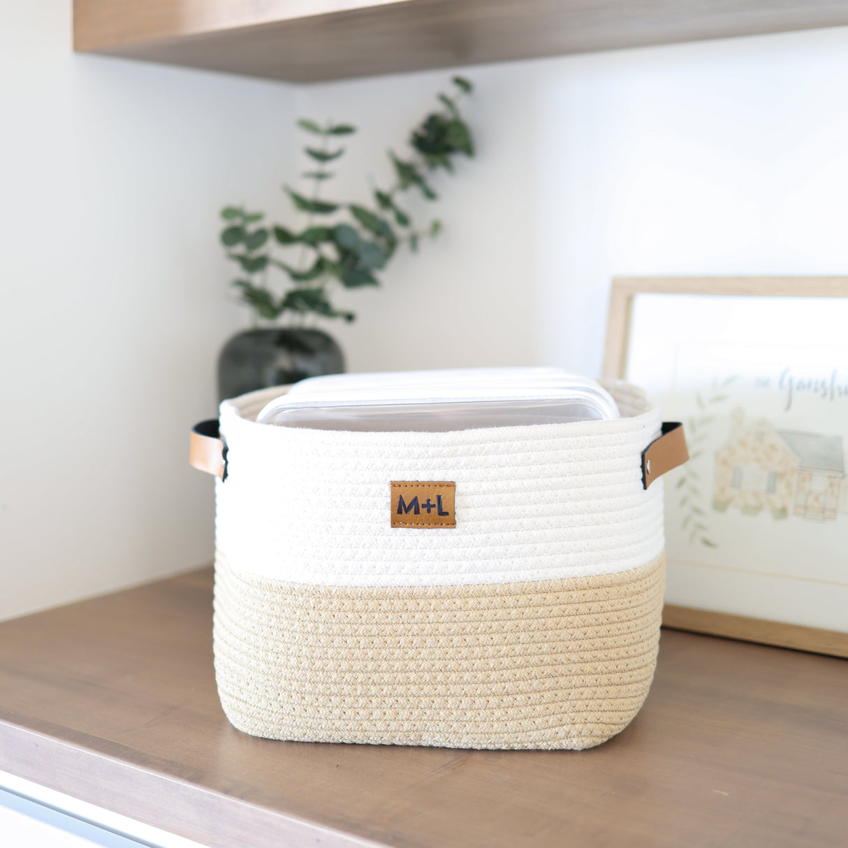 Woven storage basket with wooden handles on a shelf with a plant and picture frame in the background.