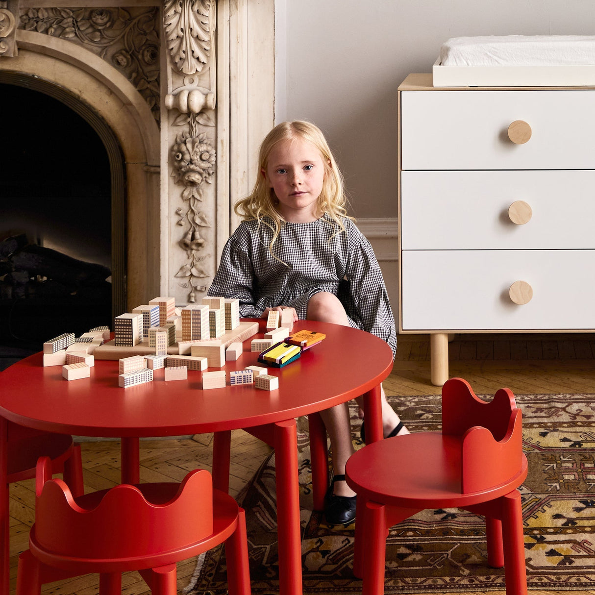 Child playing with building blocks at a red table in a room with a fireplace and white dresser.