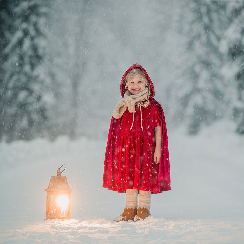 Child in a red magic cape standing in a snowy forest with a lantern
