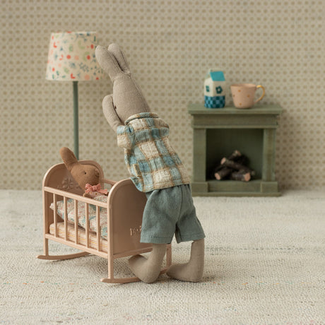 Miniature scene with a rabbit in a plaid shirt standing next to a baby crib with a teddy bear inside.