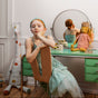 Young girl in a green dress sitting at a vanity mirror with a stuffed animal, surrounded by toys.