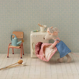 Small toy mouse in a kitchen setting with a chair, washing machine, and utensils.