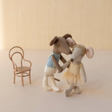 Two stuffed animal toys, one in a dress and one in a suit, standing next to a small chair on a light background.