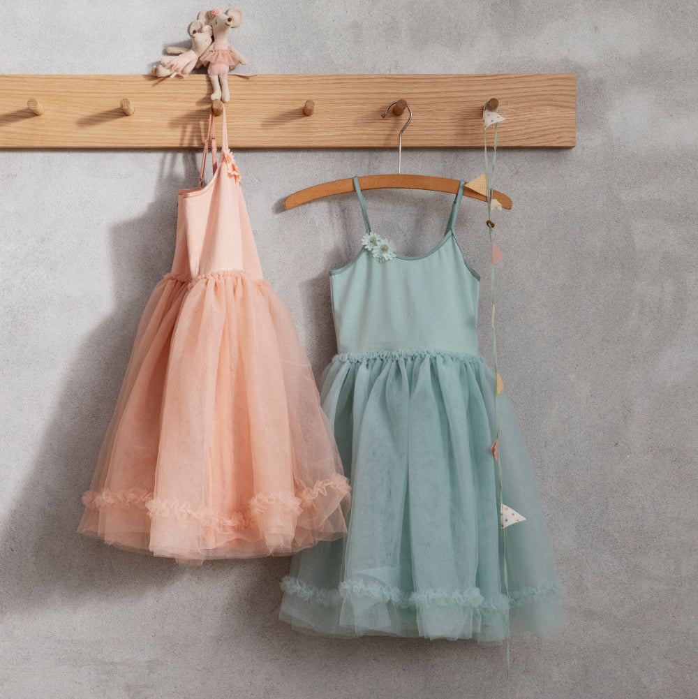 Two children's dresses, one pink and one green, hanging on a wooden rack against a gray wall.