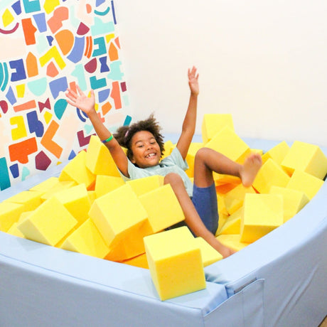 A smiling child lies on their back inside a light blue foam pit, surrounded by bright yellow foam cubes. 