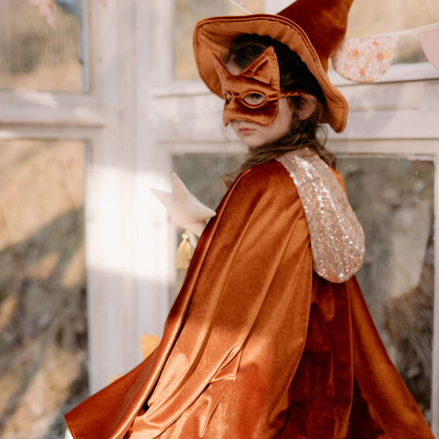 Child in a brown cape witch costume with a mask indoors.