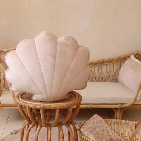 Shell-shaped cushion on a small table with a wicker chair and basket in the background.