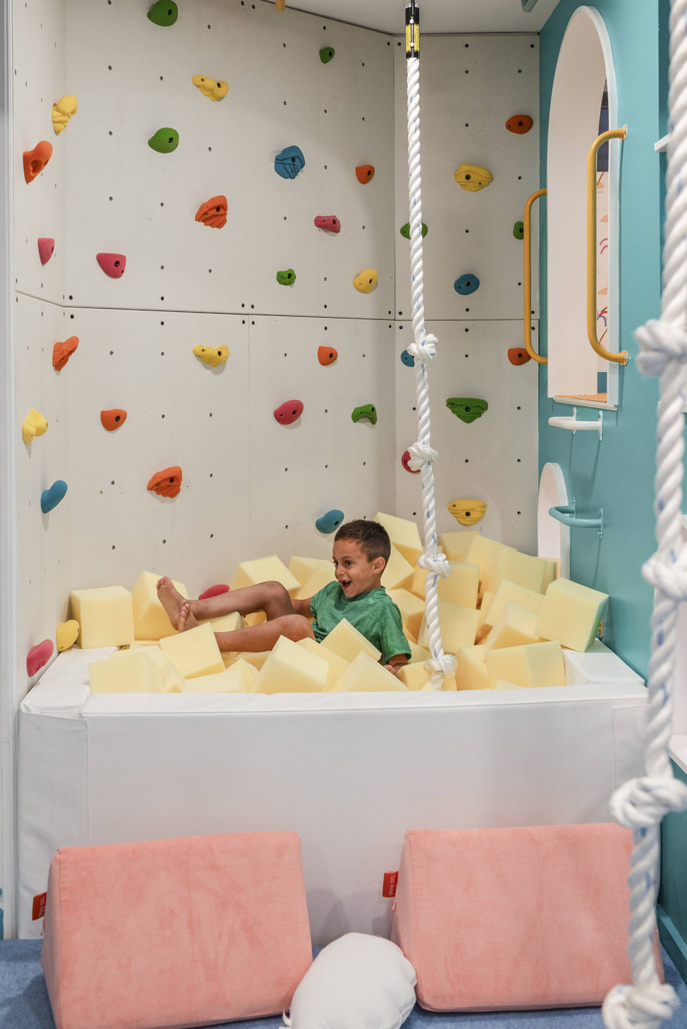 A child sits in a foam pit play area inside a playroom, surrounded by yellow foam blocks, pink floor cushions, a climbing wall, and hanging ropes.