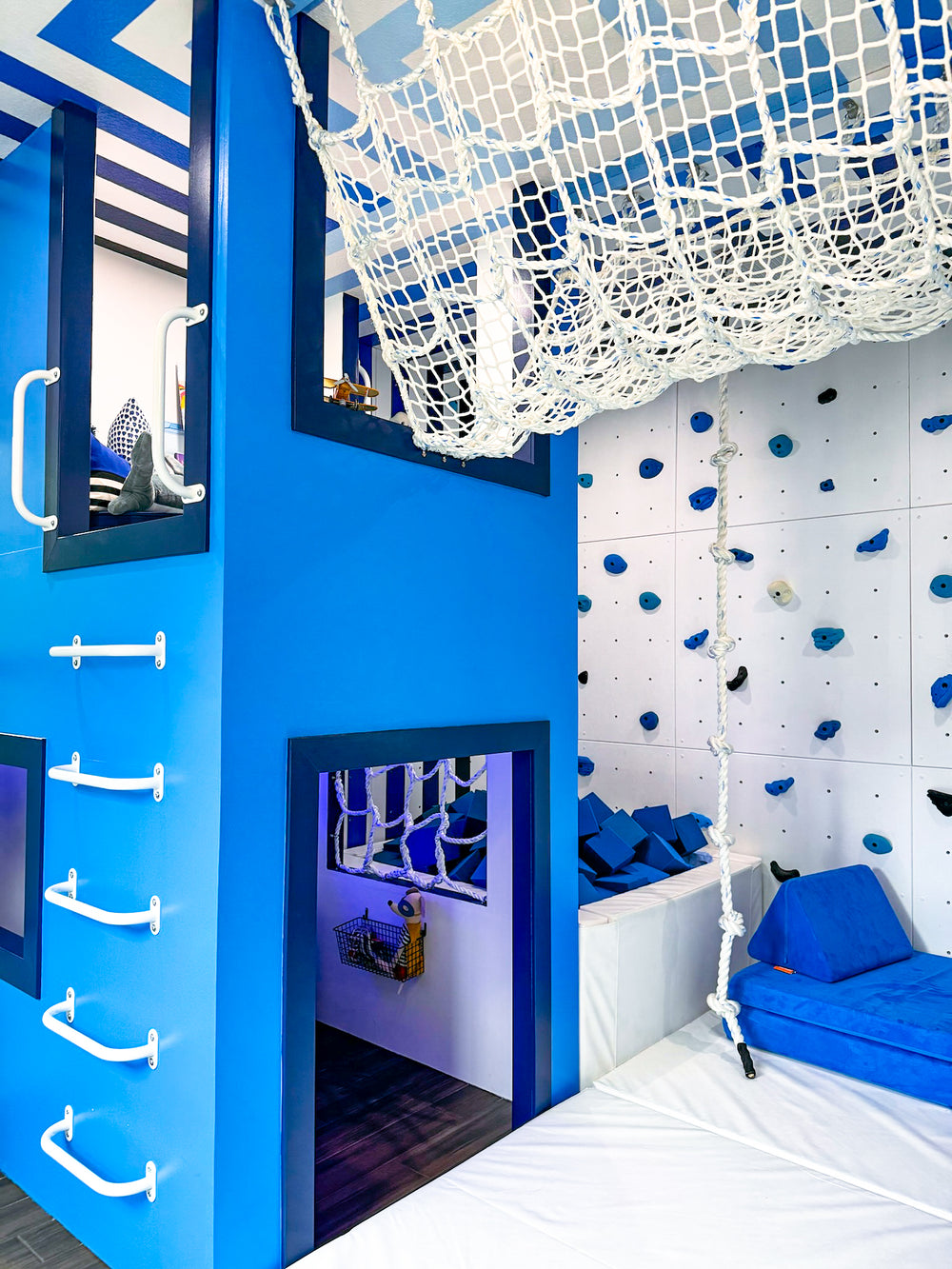 A vibrant children's playroom featuring a bright blue multi-level climbing structure with white rope handles, a white climbing wall with blue and black holds, a white hanging net, and a blue padded lounge chair on a white mat.