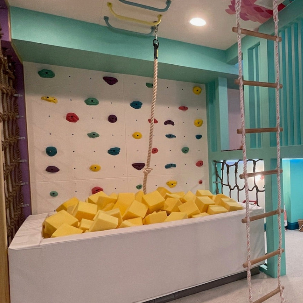 A colorful children's playroom featuring a climbing wall with multi-colored handholds, a foam pit filled with yellow blocks, a wooden climbing ladder, and yellow floor padding under a whimsical painted ceiling and wall.