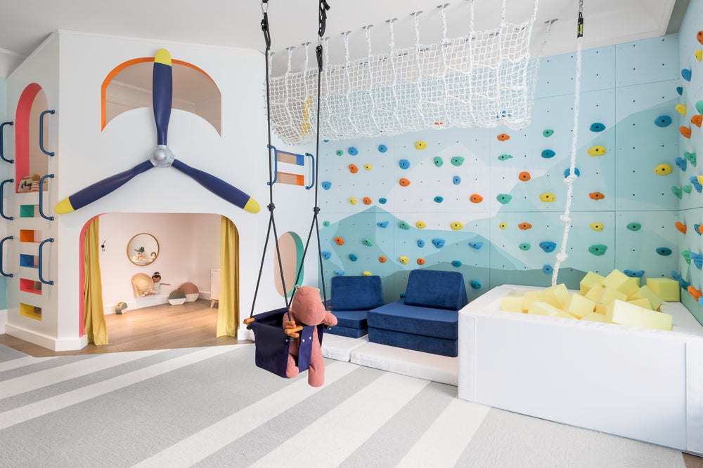 A children's indoor play area with a climbing wall, foam pit, hanging swing, net, and a white play structure shaped like an airplane cockpit. The room has a light blue wall mural, gray-and-white striped floor, and blue seating.