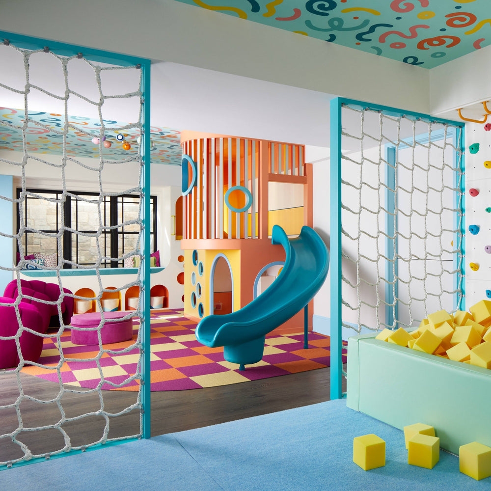 A colorful children's playroom interior with a turquoise slide, orange and yellow climbing structure, netted play doors, yellow foam blocks, and a blue carpeted floor.