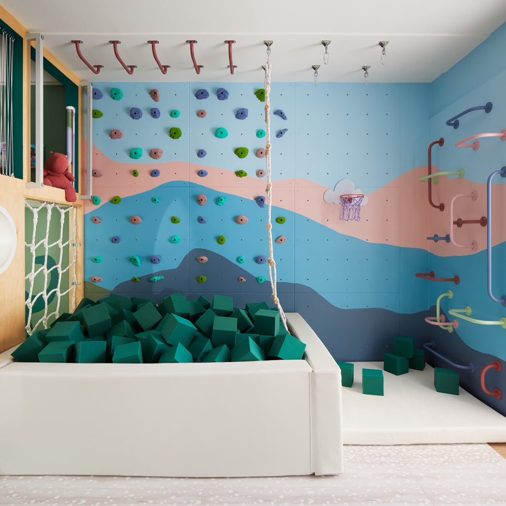 A children's indoor play area with a foam pit filled with green blocks, a climbing wall, colorful handholds, and a blue, pink, and gray mural on the wall.