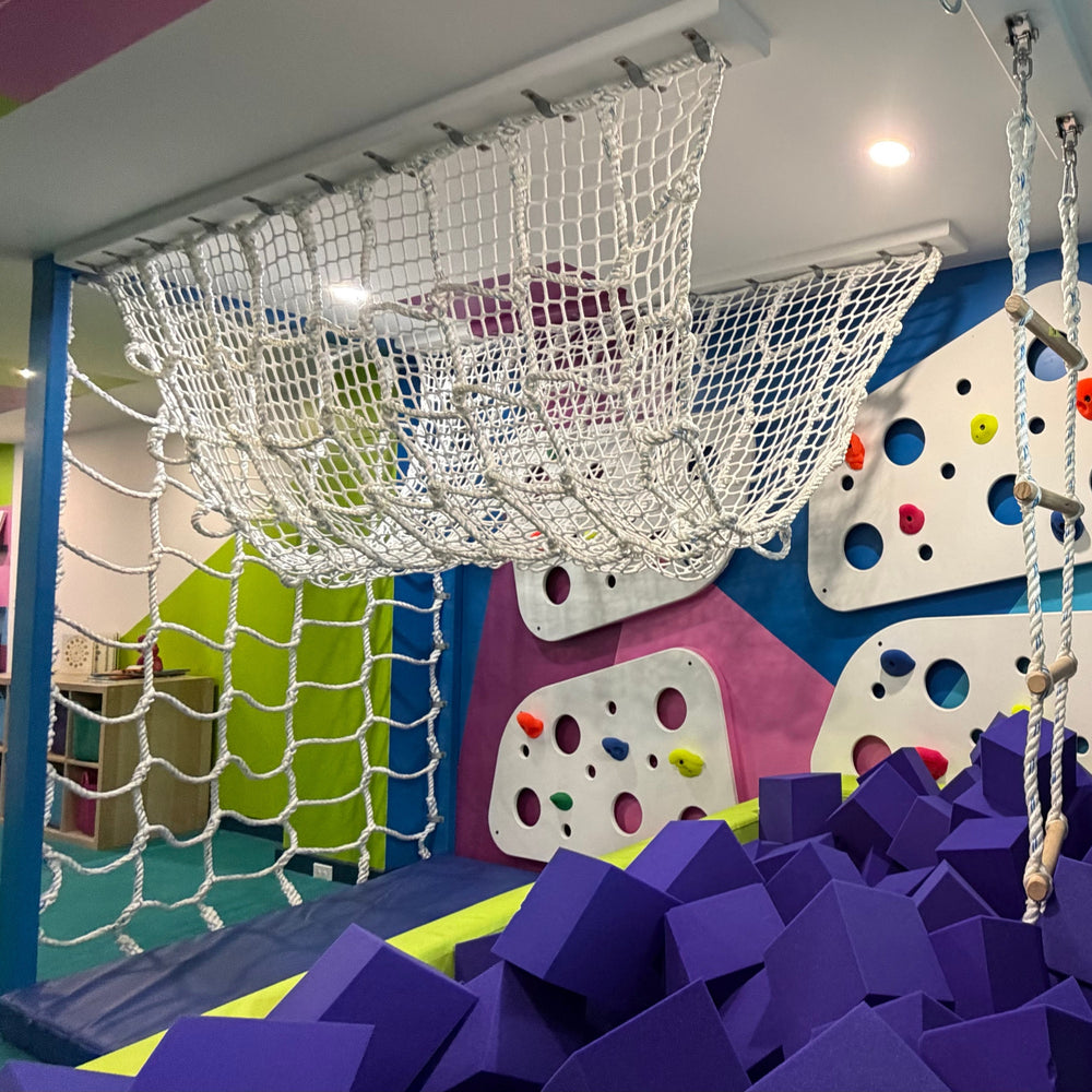 An indoor playground scene with a large white rope net hanging overhead, a foam pit filled with purple blocks in the foreground, and colorful climbing walls with white panels and multicolored holds in the background.
