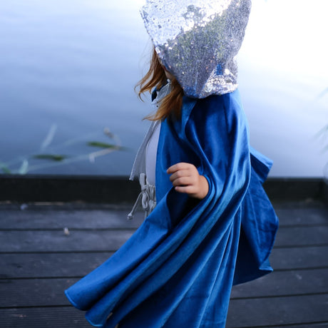Child wearing a blue magic cape and silver sequin headpiece by a body of water.