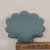 Teal shell-shaped object on a wooden shelf against a light gray wall