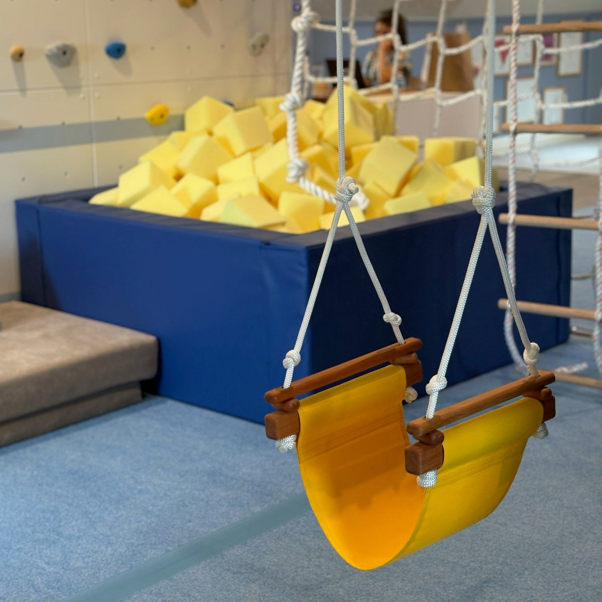 Children's swing with yellow seat in an indoor play area with soft foam blocks and climbing structures.