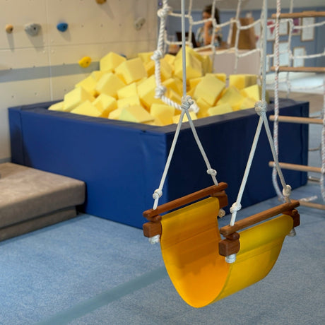 Children's swing with yellow seat in an indoor play area with soft foam blocks and climbing structures.