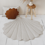 Decorative shell-shaped rug on a white wooden floor with a plant and pillow in the background.