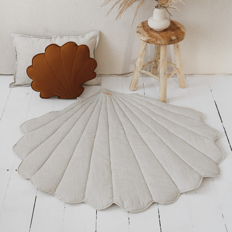 Decorative shell-shaped rug on a white wooden floor with a plant and pillow in the background.