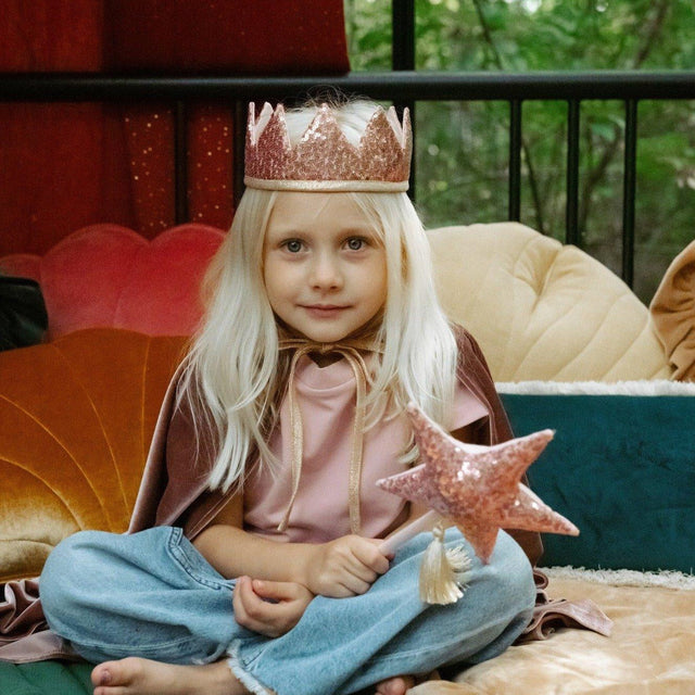 Child in costume with a star wand and pink sequins fairy tale crown, sitting on a colorful couch.