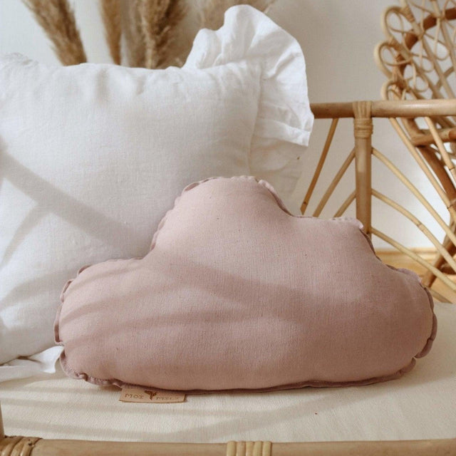 Pink cloud-shaped pillow on a wooden surface with a white pillow and decorative elements in the background.