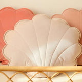 Shell-shaped cushions in pink and white on a wooden shelf against a beige wall.