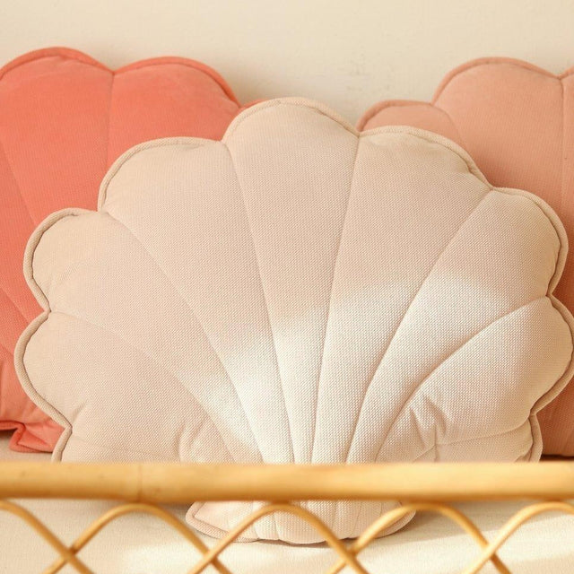 Shell-shaped cushions in pink and white on a wooden shelf against a beige wall.